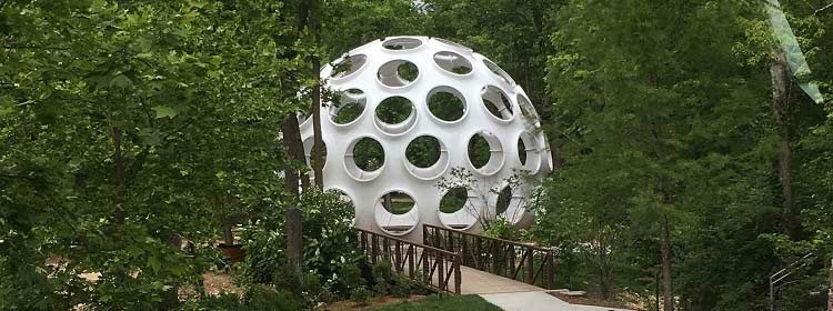 Spherical structure with circular cutouts in a forest, accessed via a wooden bridge.