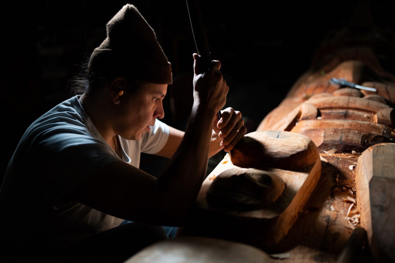 Person carving a wooden sculpture with a chisel in dim light, surrounded by wood shavings.