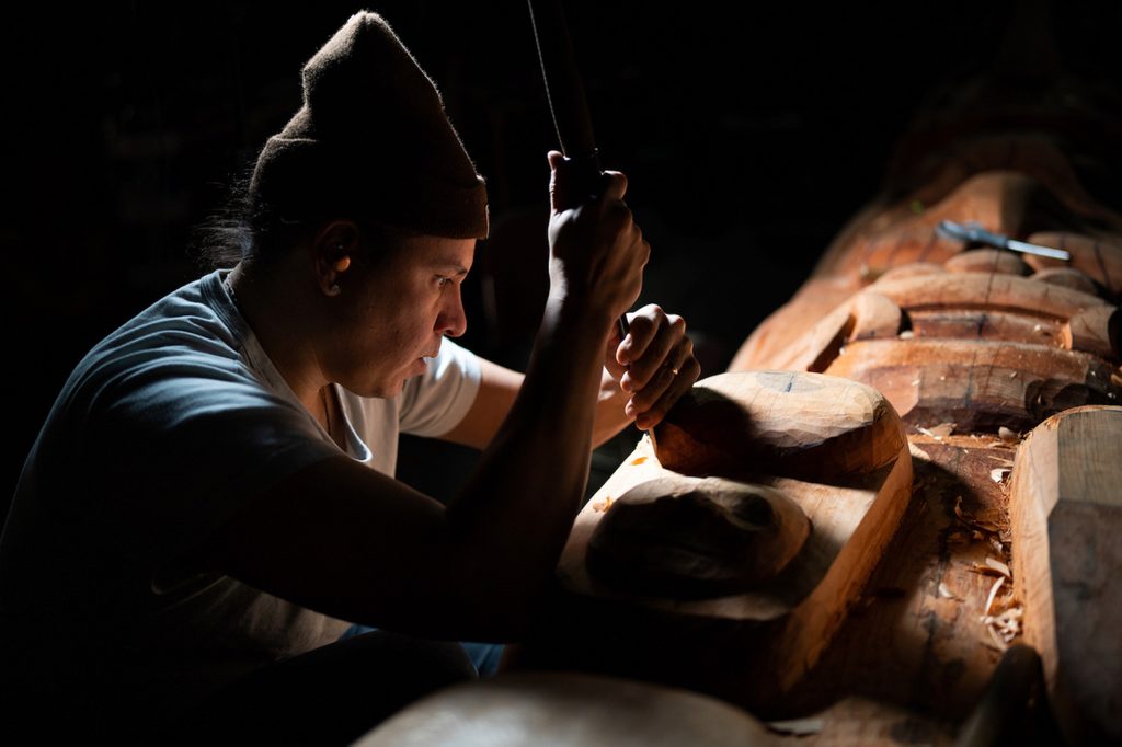 Person carving a wooden sculpture with a chisel in dim light, surrounded by wood shavings.