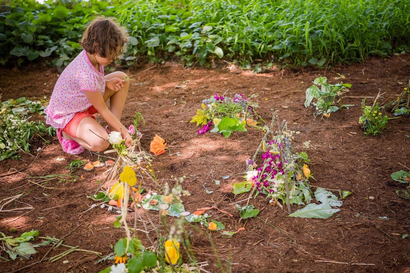 Child kneeling outdoors arranging flowers and leaves into clusters on dirt surrounded by greenery.