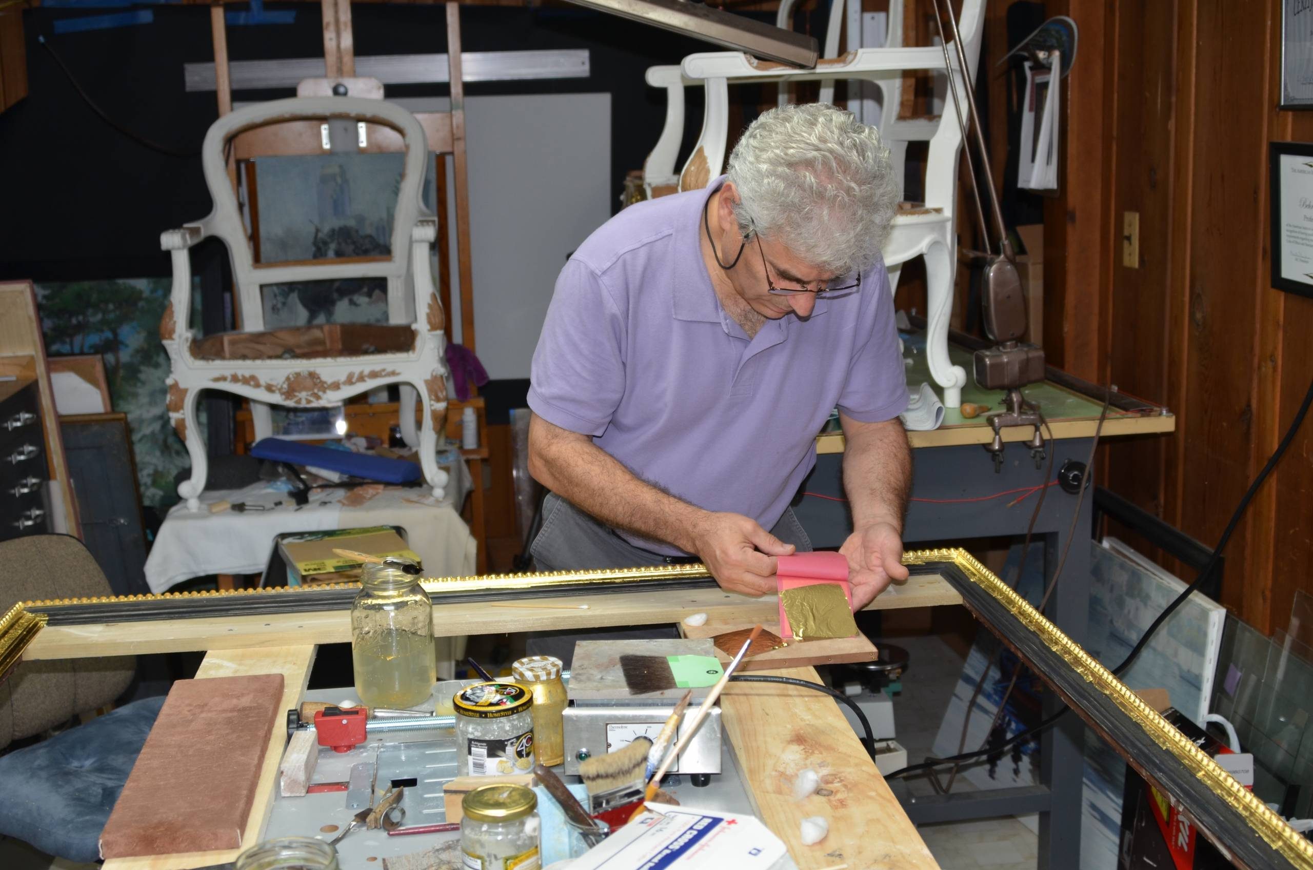 Individual crafting with gold leaf in a workshop surrounded by tools and furniture components.