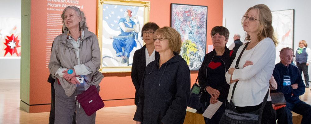 People gathered in an art gallery; a guide in plaid shirt speaks using a microphone headset.