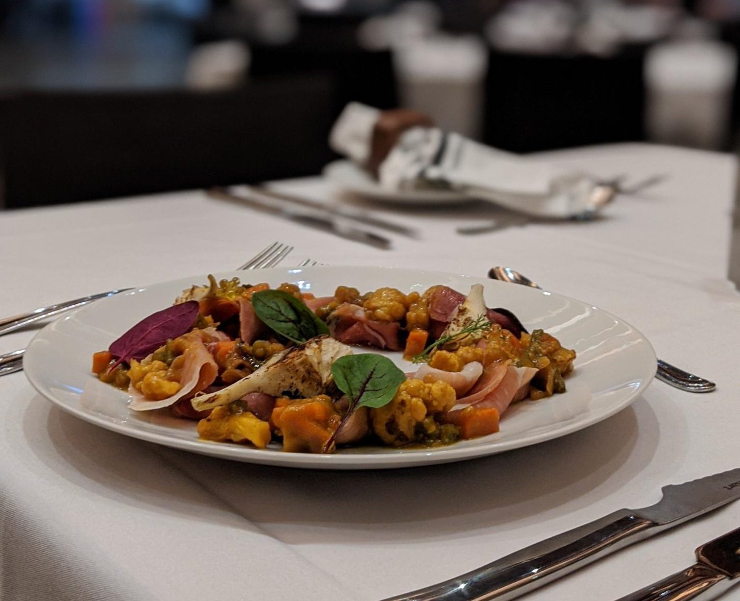 Plated dish of veggies and cured meat on a table with silverware.