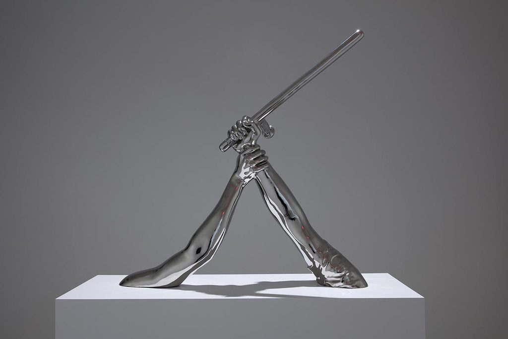 Metallic sculpture of two hands and forearms holding a sword on a pedestal.