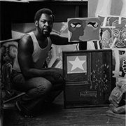 Man in studio with paintings, including abstract art with a star and facial features, sitting on floor.