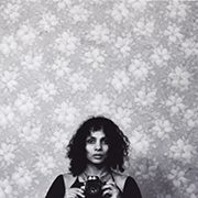 Person with curly hair holding a camera, floral wallpaper, black and white image.