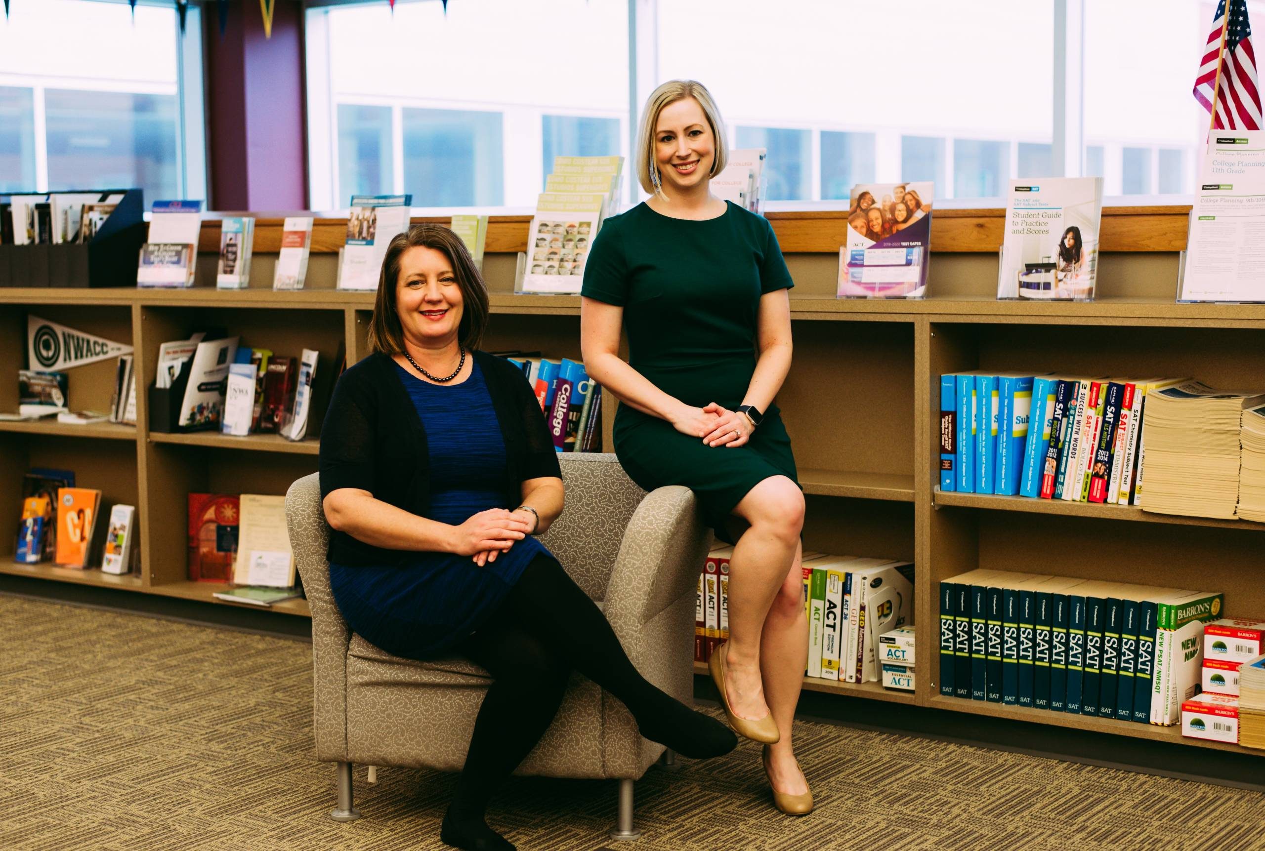 Two women in library with books, brochures, and American flag on shelf in background.