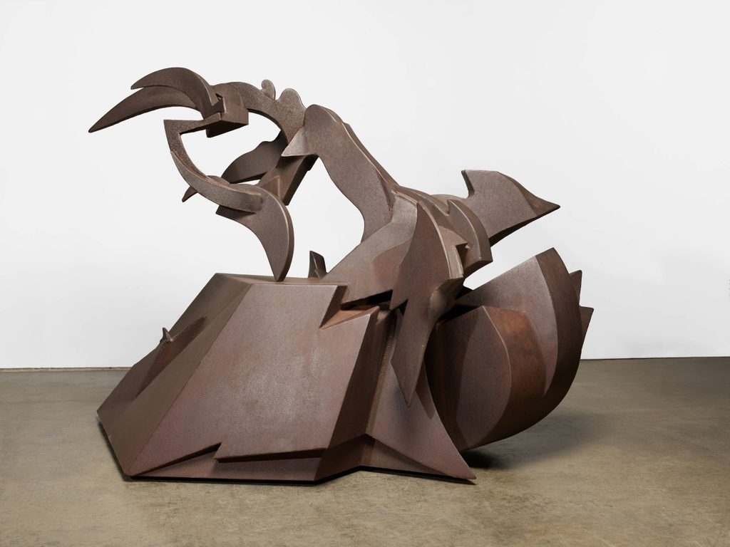 a rusted steel sculpture shaped like a train engine and wheel on the bottom with a scythe shape on top