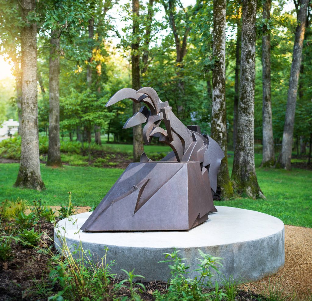 a steel gray sculpture shaped like a train engine and wheel on the bottom with a scythe shape on top sitting on a platform surrounded by a lush green forest