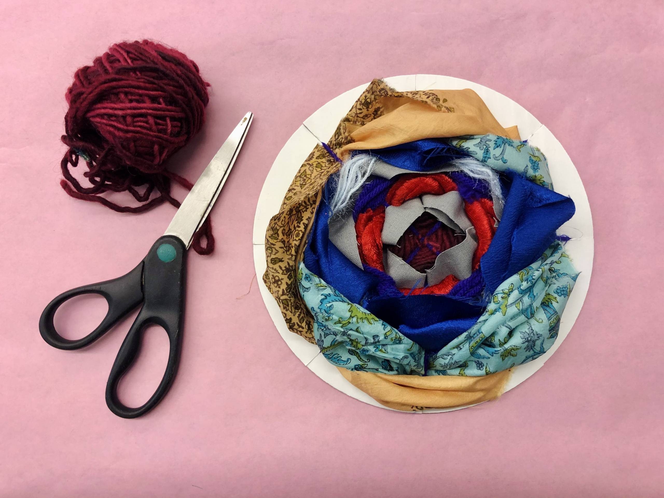 image of a weaving art activity - blue, red, and gray fabrics woven together in a circle with scissors and yarn next to it