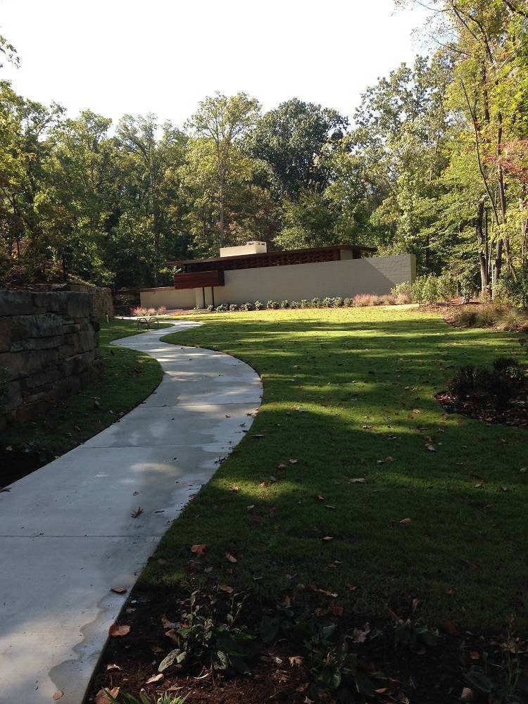 Modern minimalist building in wooded area with landscaped lawn and curved concrete path.
