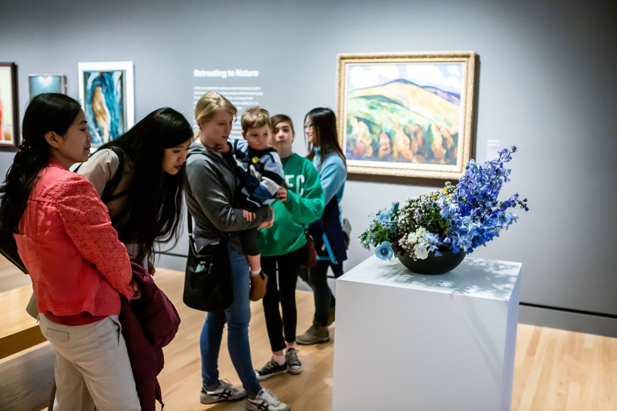 People viewing floral display on pedestal in a gallery, landscape painting in background.