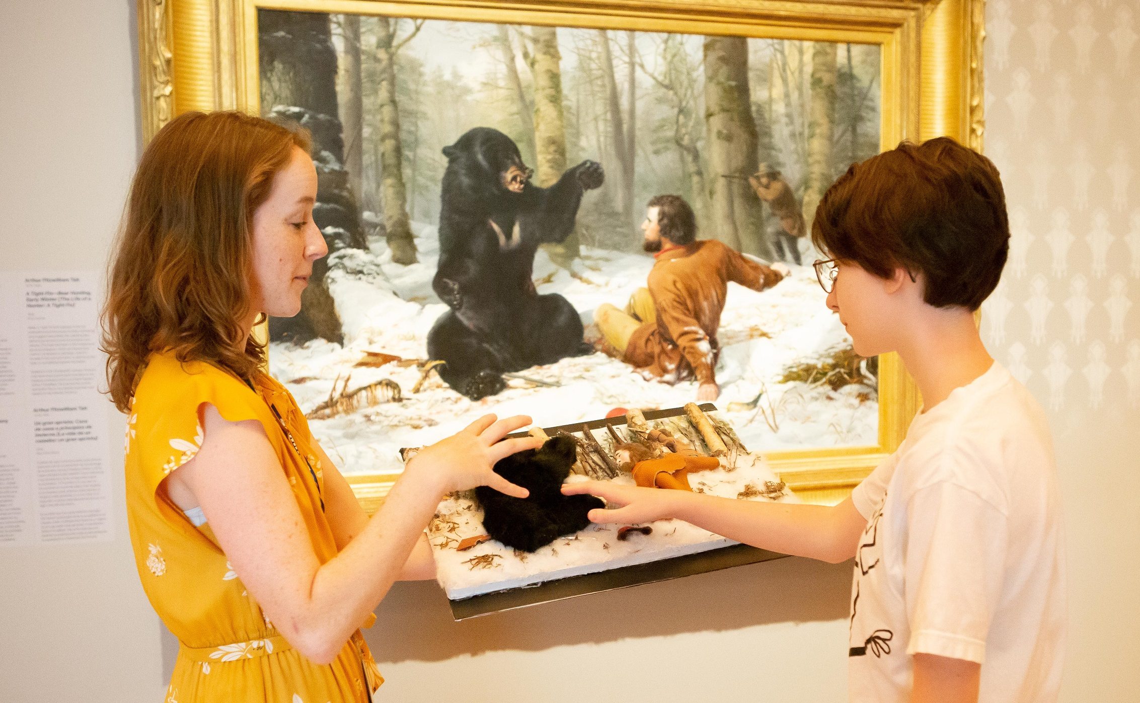 Two people inspect a tactile display with a bear figure; historical bear scene painting behind.