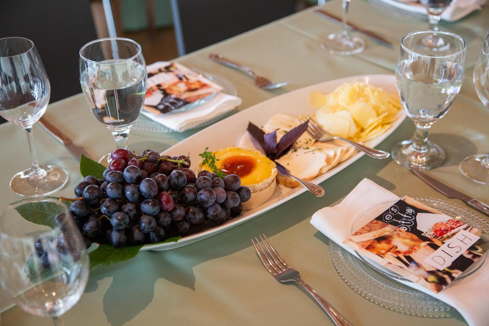 Table setting with wine glasses, cheese platter, pastry, grapes, and plates with napkins and menus.