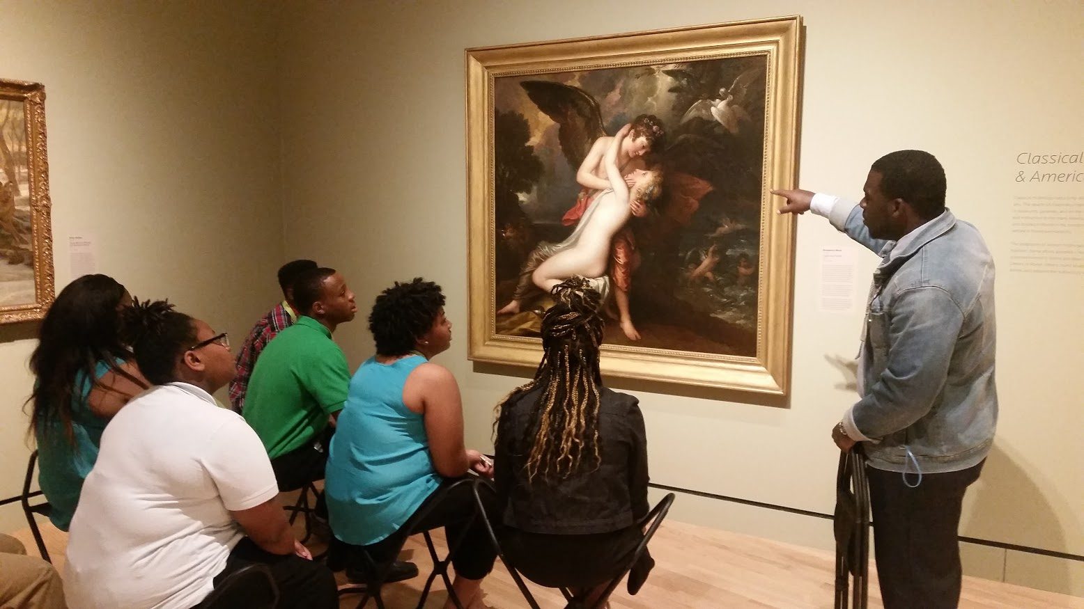 Group in museum listens to guide explaining a classical painting, pointing at details.