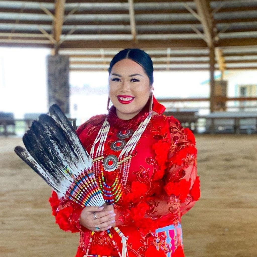 Person in traditional attire with beadwork, holding feather fan in an open, roofed structure.
