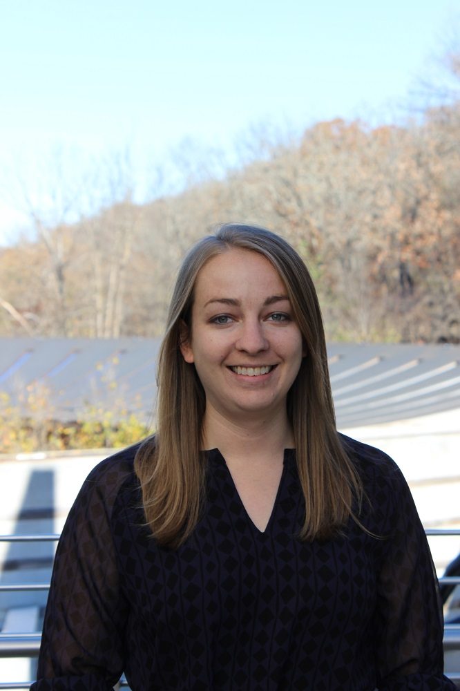Person with long hair and dark blouse smiling outdoors, bare trees and clear sky in background.