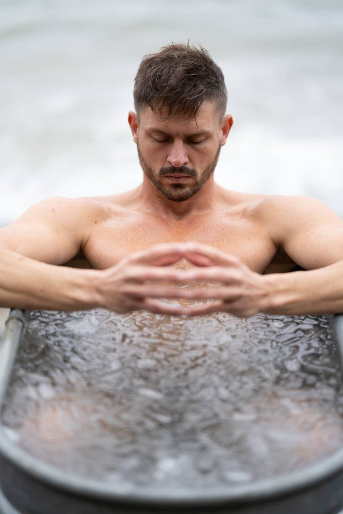 Man with closed eyes sitting shirtless in ice-filled metal tub with hands clasped.