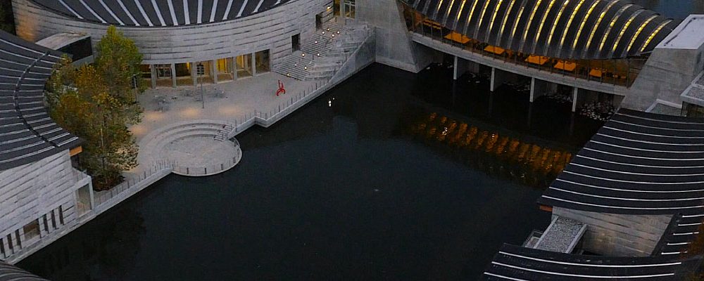 Modern curved buildings with dark roofs, glass windows, wooded autumn area, large water body.