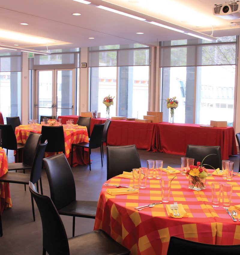 Banquet room with checkered tablecloths, set tables, black chairs, and buffet near large windows.