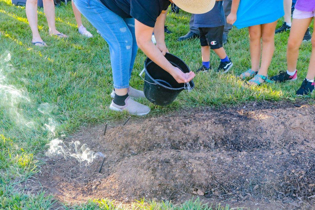 a person pours water over planted seeds with her hand as smoke billows from ceremonial sticks staked in the ground