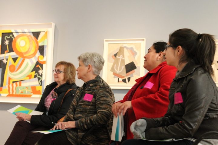 Four women with pink name tags seated, viewing framed abstract art indoors.