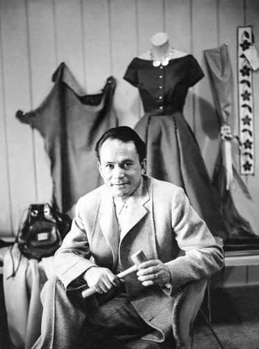 black and white photograph of fashion icon lloyd kiva new posing in front of a dress and other garments against a wall