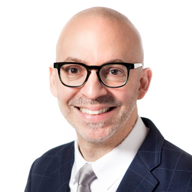 Smiling bald person with black-rimmed glasses, white shirt, dark checkered suit