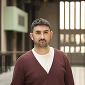 Man in burgundy sweater and white shirt standing indoors with large windows and natural light.