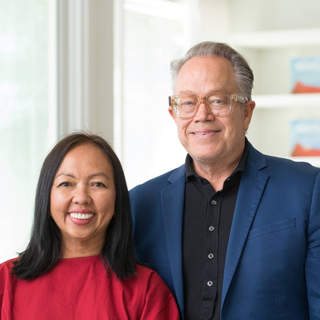 Middle-aged man and woman standing indoors near a window and bookshelf, smiling at camera.