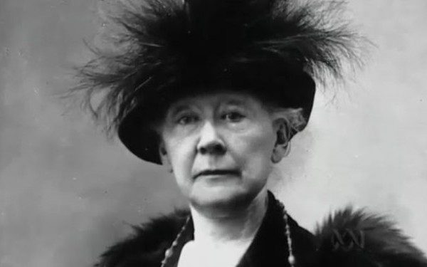 Elderly woman in black and white, wearing feathered hat and fur-trimmed coat, neutral expression.