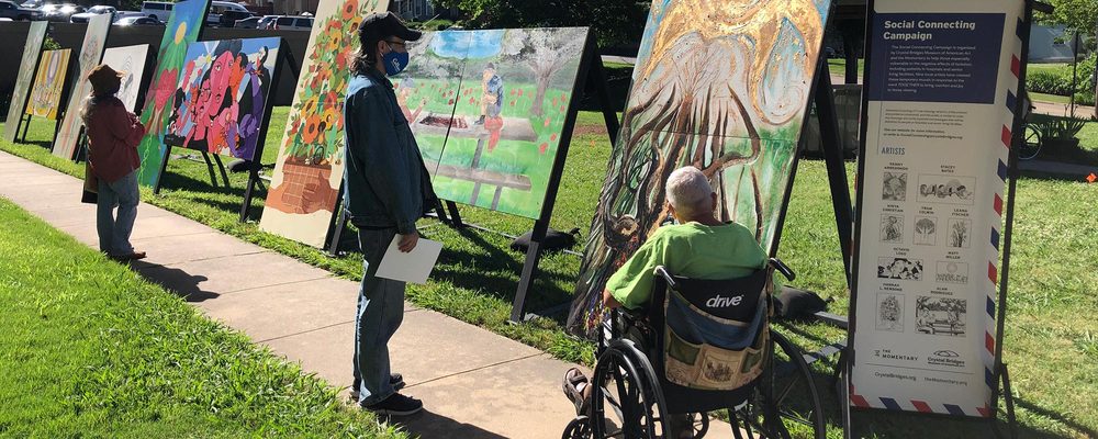 Outdoor art display with paintings on easels and viewers, including a wheelchair user, along a path.
