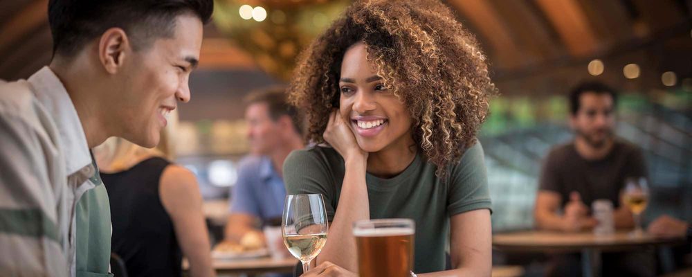 Man and woman smiling at each other, seated at a table with wine and beer, in a social setting.