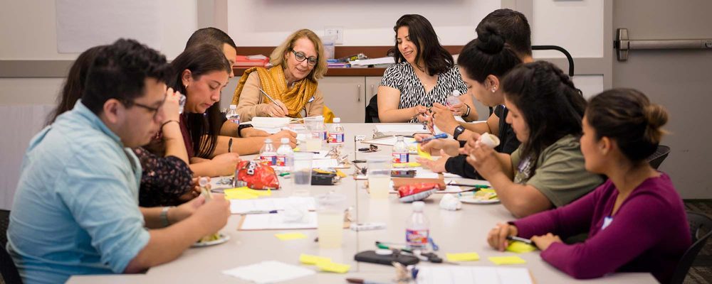 Group of people in a meeting room around a table, discussing and taking notes.