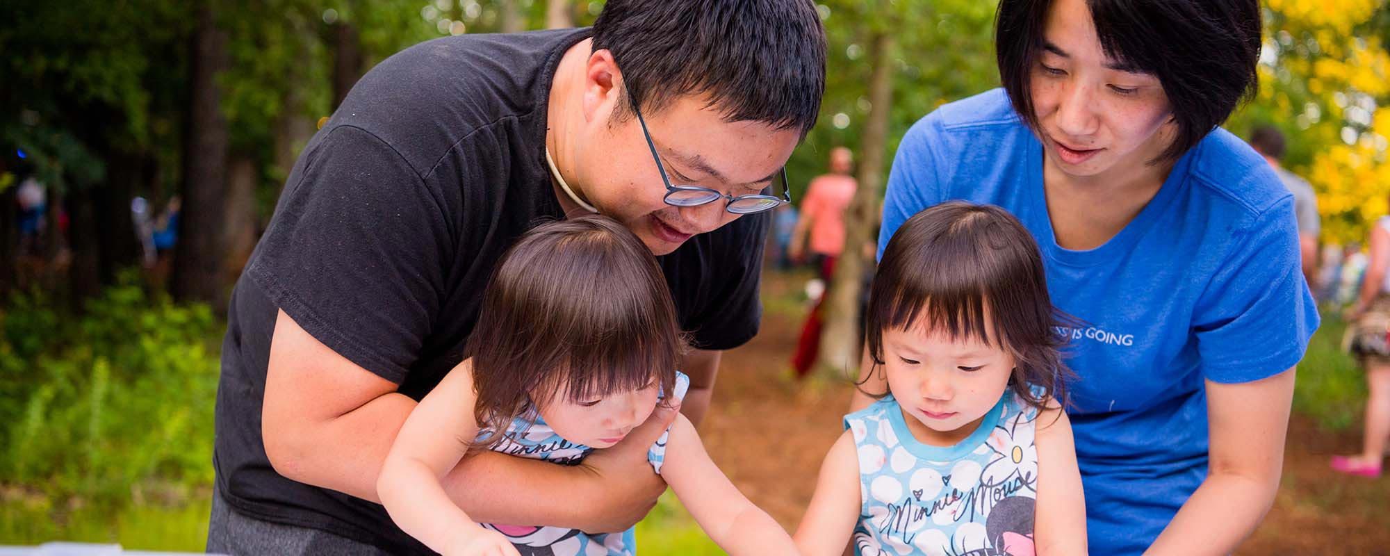 Family outdoors, two adults with smiling young children; child in Minnie Mouse shirt. Trees visible.