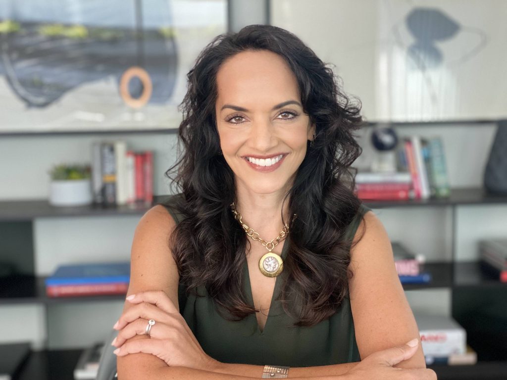Woman with dark curly hair wearing a sleeveless top and gold necklace sitting with arms crossed in front of bookshelf.