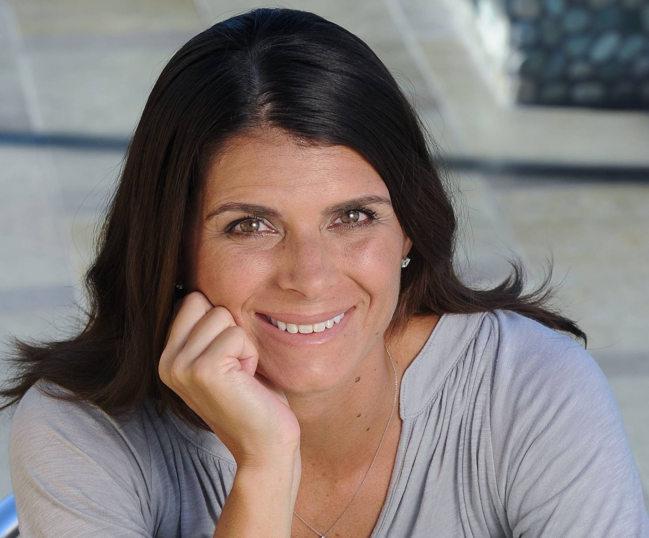 Woman with medium-length dark hair wearing a light gray top, smiling with chin resting on hand
