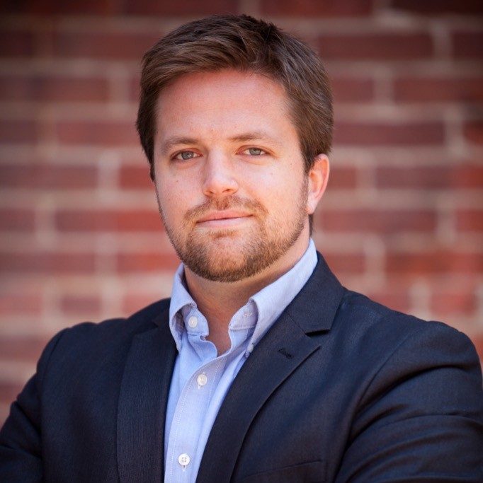 Man with short light brown hair, beard, dark suit, light blue shirt, blurred brick wall background.