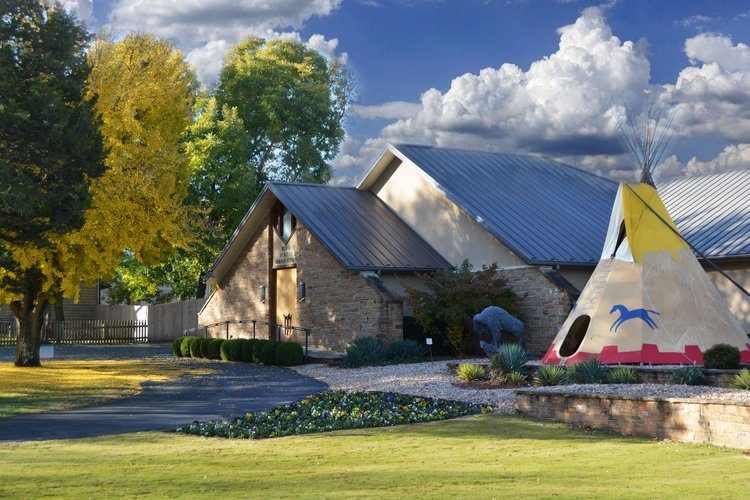 the museum of native american history exterior view with trees on the left side and a teepee and buffalo sculpture on the right side