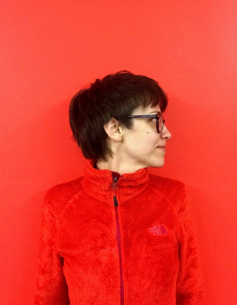 Person in glasses and red fleece jacket against a red background, looking to the side.