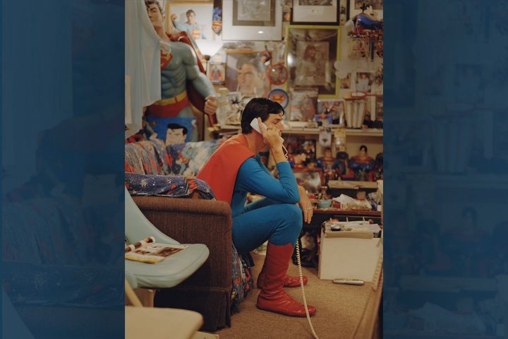 Person in Superman costume holding phone in room with Superman memorabilia, posters, and toys.