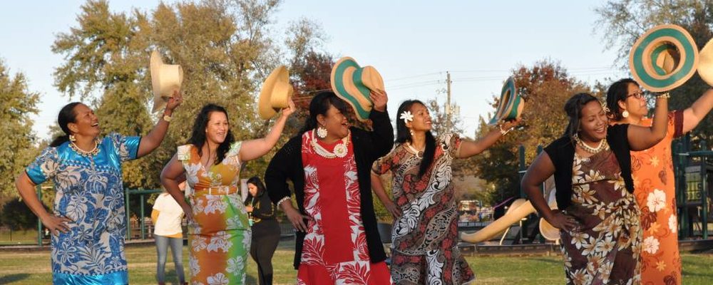 Women in colorful dresses dance outdoors, holding hats, with trees and playground in background.