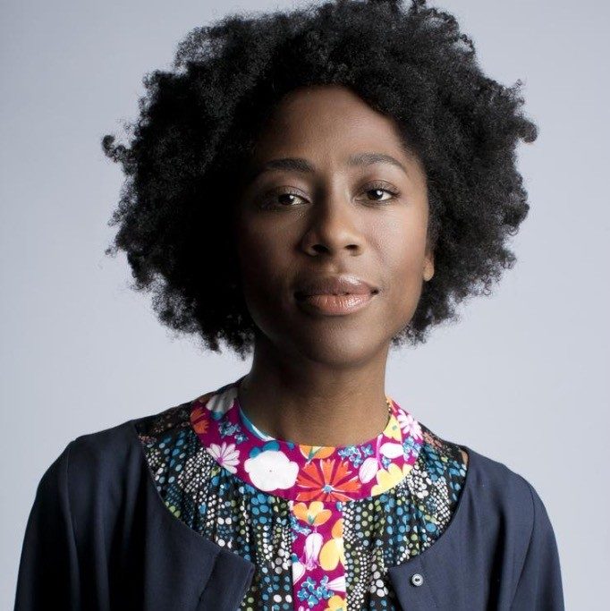 Person with curly hair in colorful top and dark jacket on plain background.