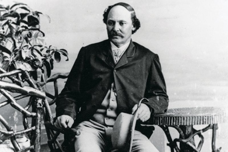 Man in suit holding hat, seated by a table with plant in background, posing for formal photo.