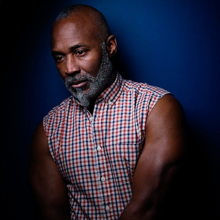 Man with beard in a sleeveless plaid shirt against a dark blue background.