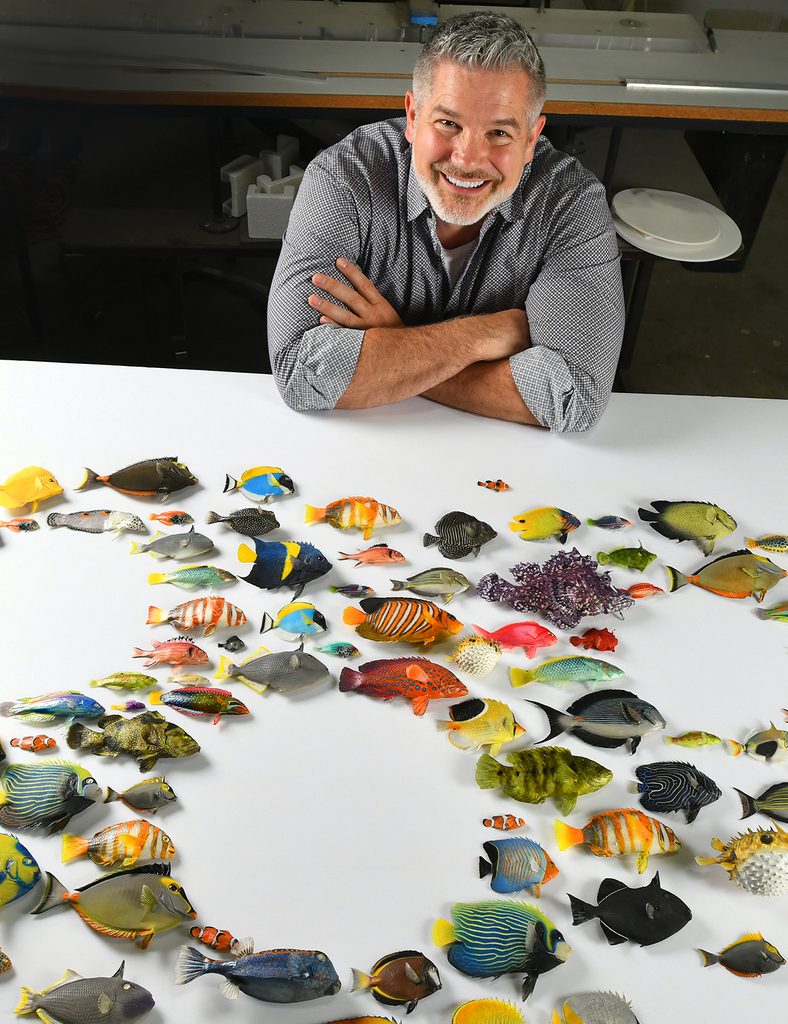 Man smiling in grey shirt sits at table with color fish mounted on a white board