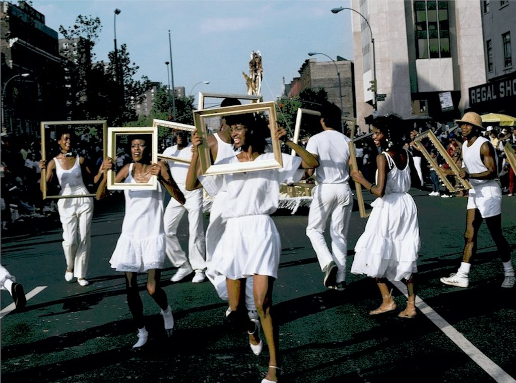 People in white outfits with wooden frames walking in a street parade with onlookers.
