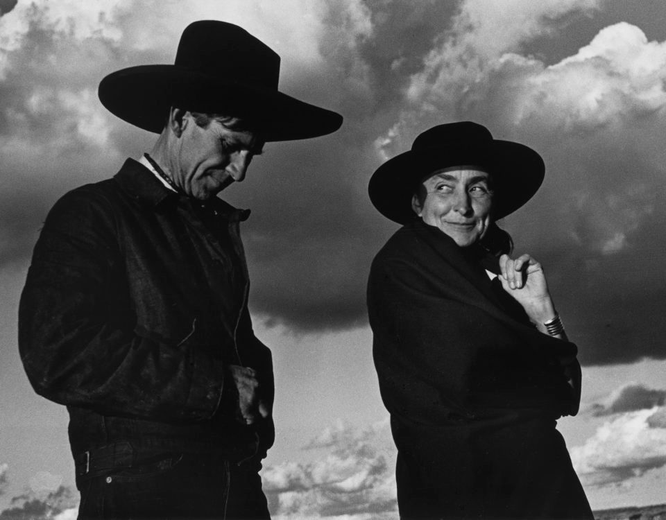 Two people in wide-brimmed hats, dark clothes, under dramatic cloudy sky, one smiles, one serious.