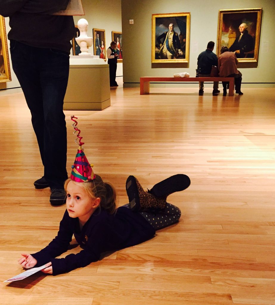 a young girl wearing a party hat lays on the floor of the gallery drawing a picture