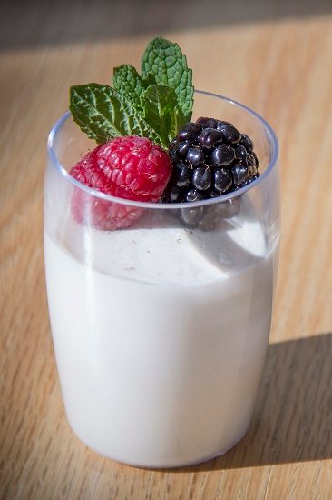 Glass cup with creamy dessert topped with raspberry, blackberry, mint on wooden surface.